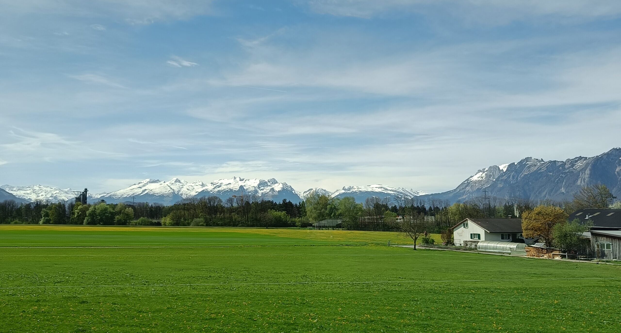 Landschaft mit Alpen im Hintergrund und zwei Häusern neben einer Wiese